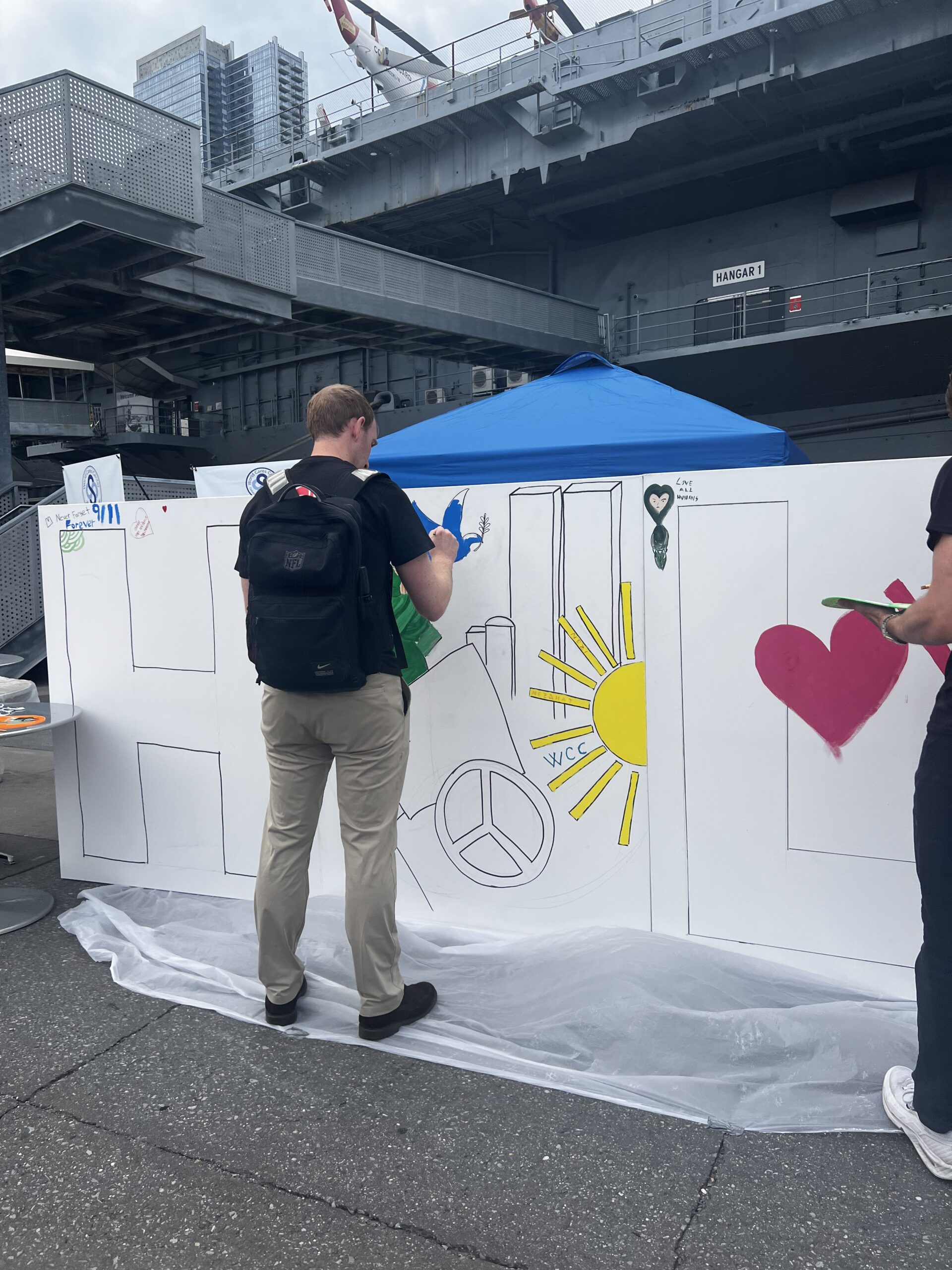 A volunteer painting a community mural with peace signs, hearts, and Never Forget 9/11 aboard the Intrepid